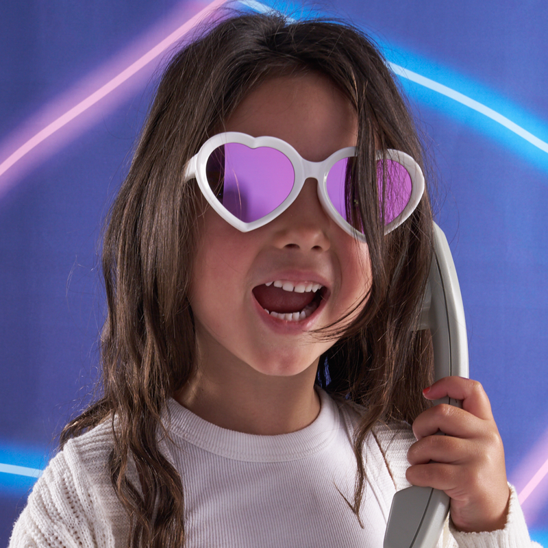 Pink | photo_of_kid.JPG | A young girl with long brown hair wearing white heart-shaped sunglasses with purple mirrored lenses, holding a gray toy phone against a blue background with neon light streaks